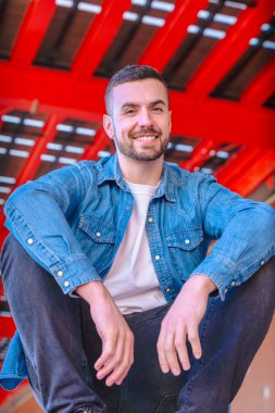 Dikey Portre gülümseyen genç adam modern kırmızı kentsel bir yapının altında gelişigüzel oturuyor. Rahat duruş, kot tarzı ve kendine güvenen bir ifade. Kameraya bakıyorum. 