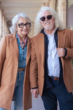 Güneş gözlüğü ve şık ceketler takan gülümseyen yaşlı bir çiftin dikey portresi. Şehir ortamında zarafet, mutluluk ve bağlantı göstererek güvenle dışarı çıkarlar.. 