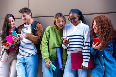 Beş çok ırklı arkadaş üniversite kampüsünde sosyal medyayı akıllı telefonlarıyla kullanırken eğleniyor. Üniversite yaşam konsepti.