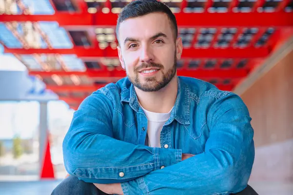 Kollarını kavuşturup kırmızı kentsel bir yapının altındaki kameraya bakan genç adam. Kendine güvenen poz, gündelik tarz ve modern, açık şehir ortamında güçlü göz teması.. 