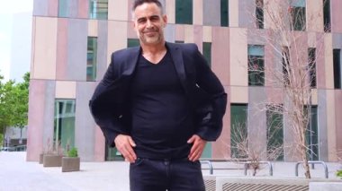 Modern bir şehir ortamında kameraya bakarken kendine güvenen siyah elbiseli olgun bir adam. Dikey binalar, gündelik pozlar ve şehir çevresindeki profesyonel varlıklar.. 