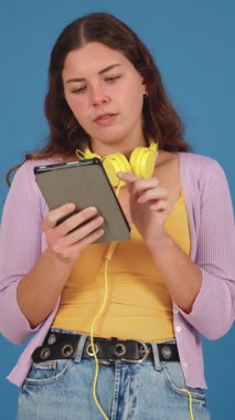 Kulaklıklı, elinde tablet olan ve düşünen genç bir kadın. Günlük kıyafetler, dikey format, mavi arka plan. Dijital ve modern bir bağlamda merak veya karar verme mekanizması.. 