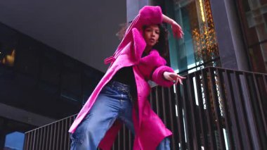 Pembe palto giyen kıvırcık saçlı zarif dansçı şehir ortamında hareketini ve enerjisini ifade ediyor. Modern moda, çağdaş dans ve sanatsal performans şehrin arka planına karşı..