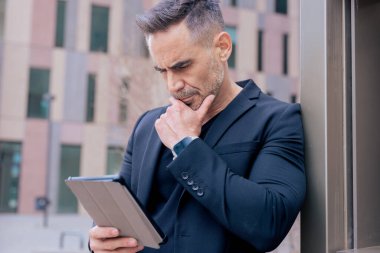 Duvara yaslanmış düşünceli bir ifadeyle tablet kullanan bir iş adamı. Şehir ortamında siyah bir takım elbise ve şık bir ayakkabı giyer. Odaklanma, planlama ve modern dijital yaşam tarzı kavramı. 