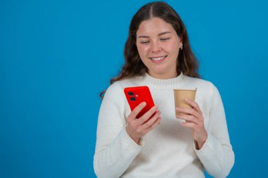 Beyaz kazaklı gülümseyen genç bir kadının portresi kırmızı bir akıllı telefon ve kahve fincanını mavi arka planda tutarak iletişimi, rahatlamayı ve gündelik modern yaşam tarzını temsil ediyor. 