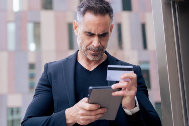 Kulaklıklı, kendine güvenen beyaz adam tablet kullanırken ve dışarıda kredi kartı tutarken ciddiydi. Çevrimiçi alışveriş kavramı, dijital ödeme, modern yaşam tarzı ve finansal teknoloji. 