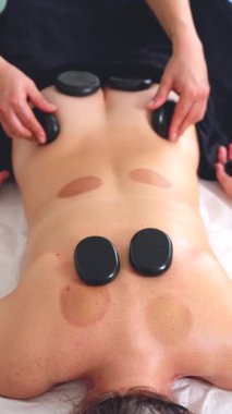 Terapist sıcak taş masajı yapıyor, müşterilerin sırtına pürüzsüz siyah taşlar sürüyor. Rahatlatıcı sağlık tedavisine yakın çekim. Spa, terapi, rahatlama ve bütünsel iyileşme kavramları için ideal.
