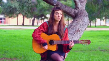 Genç bayan müzisyen, sakin bir açık hava parkında akustik gitarla şarkı söylüyor. Doğa tarafından çevrelenmiş, tutku, yaratıcılık ve duyguyu gündelik bir ortamda müzik yoluyla ifade eder.. 