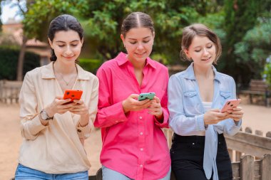 Yeşil bir parktaki ahşap bankta üç beyaz kadın, akıllı telefonlar kullanırken gülümsüyorlar. Mutlu ve meşgul görünüyorlar, modern cihazlar ve sosyal medya uygulamalarıyla açık havada eğleniyorlar.
