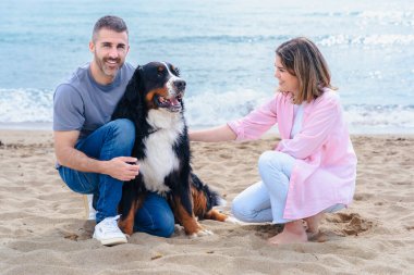 Tatil sırasında kumsalda büyük bir köpekle gülümseyen beyaz bir çiftin portresi. Deniz kenarında rahat ve keyifli bir an. Üçü de aydınlık bir günde kameraya bakıyor.. 