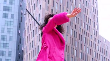 Pembe palto giyen kıvırcık saçlı zarif dansçı şehir ortamında hareketini ve enerjisini ifade ediyor. Modern moda, çağdaş dans ve sanatsal performans şehrin arka planına karşı..