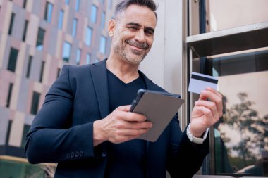 Kulaklıklı, tablet kullanırken gülümseyen ve dışarıda kredi kartı tutan kendine güvenen beyaz bir adam. Çevrimiçi alışveriş kavramı, dijital ödeme, modern yaşam tarzı ve finansal teknoloji. 