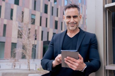 Kendine güveni tam bir işadamı modern şehir ortamında tablet kullanırken gülümsüyor. Siyah bir takım elbise ve şık bir elbise giyiyor. Başarı kavramı, teknoloji, dijital yaşam tarzı ve üretkenlik. Boşluğu kopyala