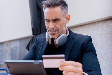 Kulaklıklı, tablet kullanırken gülümseyen ve dışarıda kredi kartı tutan kendine güvenen beyaz bir adam. Çevrimiçi alışveriş kavramı, dijital ödeme, modern yaşam tarzı ve finansal teknoloji. 