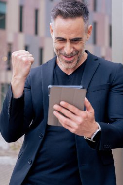 Kendine güvenen işadamı online bahsi kazanmasını kutluyor, elinde tablet, yumruğunu kaldırıyor. Şehir ortamında açık havada gülümsüyor. Başarı kavramı, dijital yaşam tarzı ve modern finans. Dikey