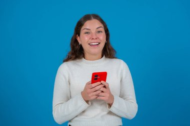 Beyaz kazaklı neşeli genç bir kadın elinde kırmızı bir akıllı telefon tutarken kameraya bakıyor, canlı mavi bir arka plana karşı duruyor, bağlantıyı, neşeyi ve modern iletişimi temsil ediyor.. 