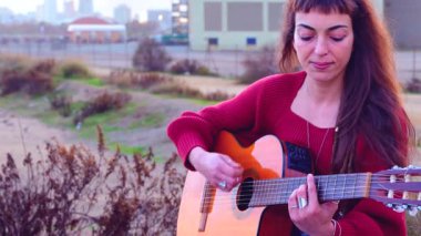 Genç bayan müzisyen, sakin bir açık hava parkında akustik gitarla şarkı söylüyor. Doğa tarafından çevrelenmiş, tutku, yaratıcılık ve duyguyu gündelik bir ortamda müzik yoluyla ifade eder.. 