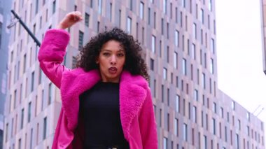 Pembe palto giyen kıvırcık saçlı zarif dansçı şehir ortamında hareketini ve enerjisini ifade ediyor. Modern moda, çağdaş dans ve sanatsal performans şehrin arka planına karşı..