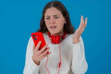 Beyaz kadın akıllı telefonundan kötü haberler okurken şok olmuş görünüyor. Kulaklık takıyor ve beyaz bir kazak giyiyor. Katı mavi arka plana karşı kaygıyla tepki veriyor..