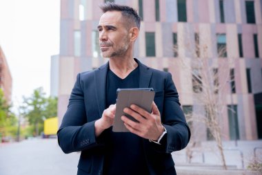 İş adamı, modern bir şehirde gözünü kaçırırken açık havada bir tablet kullanıyor. Siyah bir takım elbise ve şık bir bant giyiyor. Teknoloji, odaklanma, hareketlilik ve dijital mesleki yaşam tarzı kavramı. 