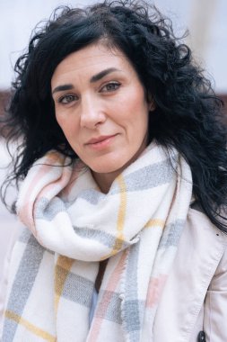 Dışarıdaki bankta oturan bir kadının dikey portresi. Bej bir ceket ve eşarp giyiyor, sakin ve kendinden emin görünüyor. Kış atmosferi ve mevsimsel moda kavramı olan şehir arkaplanı. 