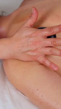 Terapist sıcak taş masajı yapıyor, müşterilerin sırtına pürüzsüz siyah taşlar sürüyor. Rahatlatıcı sağlık tedavisine yakın çekim. Spa, terapi, rahatlama ve bütünsel iyileşme kavramları için ideal.