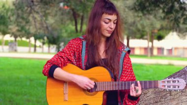Genç bayan müzisyen, sakin bir açık hava parkında akustik gitarla şarkı söylüyor. Doğa tarafından çevrelenmiş, tutku, yaratıcılık ve duyguyu gündelik bir ortamda müzik yoluyla ifade eder.. 
