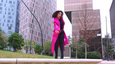 Pembe palto giyen kıvırcık saçlı zarif dansçı şehir ortamında hareketini ve enerjisini ifade ediyor. Modern moda, çağdaş dans ve sanatsal performans şehrin arka planına karşı..