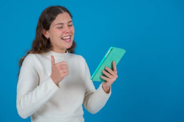 Tablet tutan ve neşeli bir ifadeyle baş parmak gösteren mutlu beyaz kadın. Fotokopi alanı olan mavi arkaplan üzerine stüdyo çekimi, iletişim ya da teknoloji kavramları için ideal. 