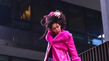 Pembe palto giyen kıvırcık saçlı zarif dansçı şehir ortamında hareketini ve enerjisini ifade ediyor. Modern moda, çağdaş dans ve sanatsal performans şehrin arka planına karşı..