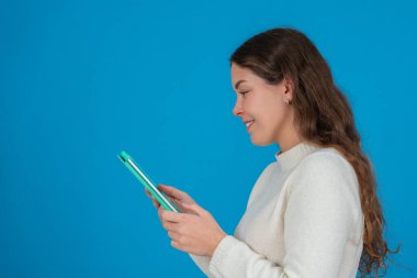 Beyaz kazak giyen gülümseyen genç bir kadının yeşil tablet kullanarak fotokopi alanındaki mavi arka plana bakması dijital iletişimi, taramayı ve teknolojik etkileşimi sembolize ediyor.. 