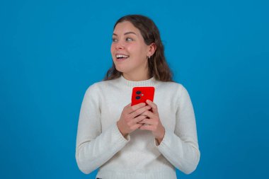 Şaşırmış bir ifadeyle yan tarafa bakarken elinde kırmızı bir akıllı telefon tutan mutlu genç kadın. Parlak mavi bir arka planın önünde duruyor, heyecan ve merakını ifade ediyor.. 