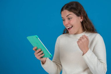 Genç ve mutlu bir kadın elinde bir tablet tutuyor ve iyi haberler aldıktan sonra sevinçle kutluyor. Heyecanla gülümsüyor ve mavi bir arka plana karşı zafer jesti olarak yumruğunu sıkıyor. Boşluğu kopyala. 