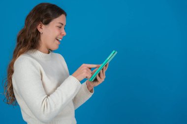 Beyaz kazak giyen gülümseyen genç bir kadının yeşil tablet kullanarak fotokopi alanındaki mavi arka plana bakması dijital iletişimi, taramayı ve teknolojik etkileşimi sembolize ediyor.. 