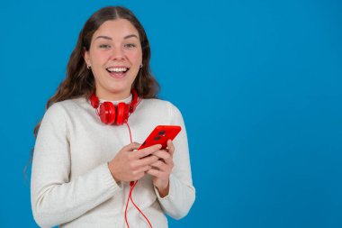 Boynuna kırmızı kulaklık takan genç bir kadın akıllı telefonuna bakıp gülümsüyor. Mutlu ve heyecanlı görünüyor, muhtemelen bir mesaja veya içeriğe tepki veriyor. Kameraya bakıyor, mavi arka plan..
