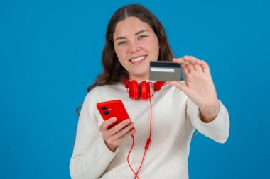 Beyaz kazaklı ve kırmızı kulaklıklı mutlu genç bir kadın kameraya bir kredi kartı gösterirken gülümsüyor ve elinde kırmızı bir akıllı telefon tutuyor, mavi arka planda online alışverişi sembolize ediyor.. 