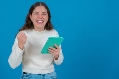  Beyaz kazaklı genç bir kadın yeşil bir tablet tutarken yumruğunu kaldırıyor ve kameraya gülümsüyor, mavi arka planda, başarıyı, neşeyi ve dijital hayatı simgeliyor. Boşluğu kopyala