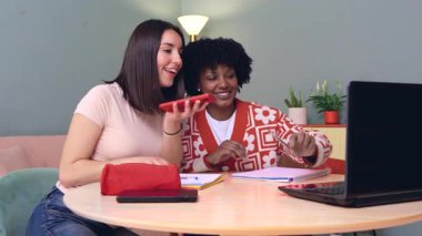 Birlikte çalışırken bilgisayar ve akıllı telefon kullanarak masada oturan gülümseyen öğrenciler. Samimi ve dost canlısı bir öğrenme alanında bir video çağrısına ya da çevrimiçi aktiviteye katılıyorlar.. 