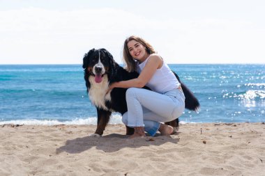 Kumsalda köpeğinin yanında diz çökmüş neşeli genç bir kadının portresi. Güneşli bir günde her ikisi de kameraya bakar, deniz kenarında huzurlu bir anın tadını çıkarırlar neşe, dostluk ve doğa ile.. 