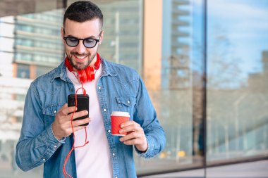 Şehir ortamında akıllı telefonunu kullanırken kırmızı kulaklıklı genç adam kahvenin tadını çıkarıyor. O okurken veya mesajlaşırken gülümsüyor, modern cam binaların önünde sıradan bir anda duruyor.. 