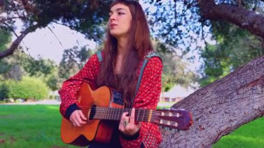 Genç bayan müzisyen, sakin bir açık hava parkında akustik gitarla şarkı söylüyor. Doğa tarafından çevrelenmiş, tutku, yaratıcılık ve duyguyu gündelik bir ortamda müzik yoluyla ifade eder.. 