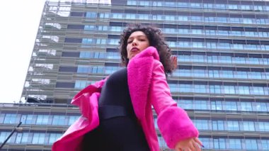 Pembe palto giyen kıvırcık saçlı zarif dansçı şehir ortamında hareketini ve enerjisini ifade ediyor. Modern moda, çağdaş dans ve sanatsal performans şehrin arka planına karşı..