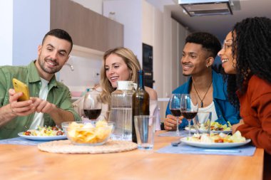 Bir grup mutlu çok ırklı arkadaş sıcak, modern bir mutfakta abur cubur ve içecek paylaşıyorlar. Parlak iç mekan, neşeli ifadeler, günlük kıyafetler, rahat ve dost canlısı sosyal atmosfer.. 