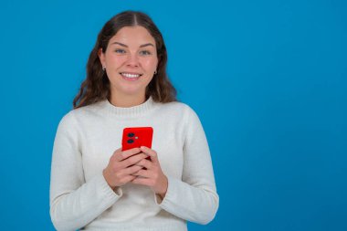 Beyaz kazaklı neşeli genç bir kadın elinde kırmızı bir akıllı telefon tutarken kameraya bakıyor, canlı mavi bir arka plana karşı duruyor, bağlantıyı temsil ediyor, neşe verici. Kopyalama alanı.