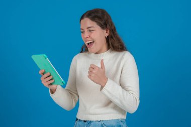 Tablet tutan ve neşeli bir ifadeyle baş parmak gösteren mutlu beyaz kadın. Fotokopi alanı olan mavi arkaplan üzerine stüdyo çekimi, iletişim ya da teknoloji kavramları için ideal. 