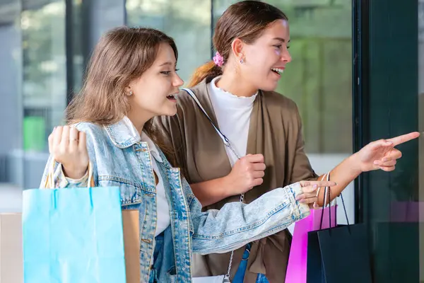 Alışveriş torbaları tutan ve vitrinlere bakan iki mutlu beyaz kadın. Şehirde alışveriş ve arkadaşlığın olduğu bir günde samimi ve neşeli bir şehir anı. 