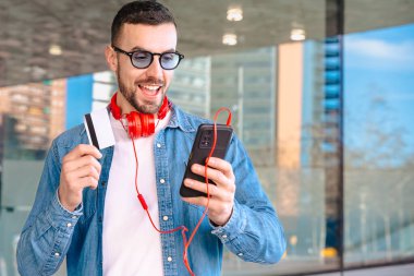 Elinde bir kredi kartı ve akıllı telefon tutarak başarılı bir online alışveriş kutlaması yapan mutlu bir adam. Modern bir şehir ortamında, kulaklık ve güneş gözlüğü takarak heyecanla gülümsüyor.. 