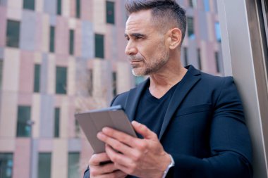 İş adamı, modern bir şehirde gözünü kaçırırken açık havada bir tablet kullanıyor. Siyah bir takım elbise ve şık bir bant giyiyor. Teknoloji, odaklanma, hareketlilik ve dijital mesleki yaşam tarzı kavramı. 