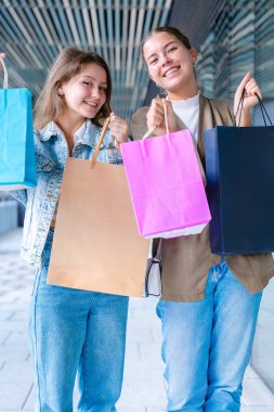 Dikey portre İki neşeli beyaz kadın kameraya bakıyor ve renkli alışveriş torbaları tutarken gülümsüyor. Açık hava ortamında neşeli ve eğlenceli alışveriş anları yakalıyorum.. 