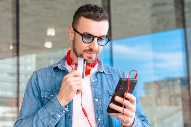 Elinde bir kredi kartı ve akıllı telefon tutarak başarılı bir online alışveriş kutlaması yapan mutlu bir adam. Modern bir şehir ortamında, kulaklık ve güneş gözlüğü takarak heyecanla gülümsüyor.. 